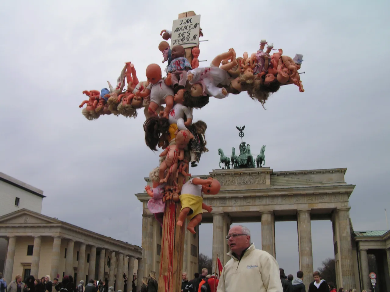 Ein Holzkreuz, über und über mit Puppen behängt, vor dem Brandenburger Tor in Berlin. Oben ein Schild mit der Aufschrift Im Namen des Herrn.
