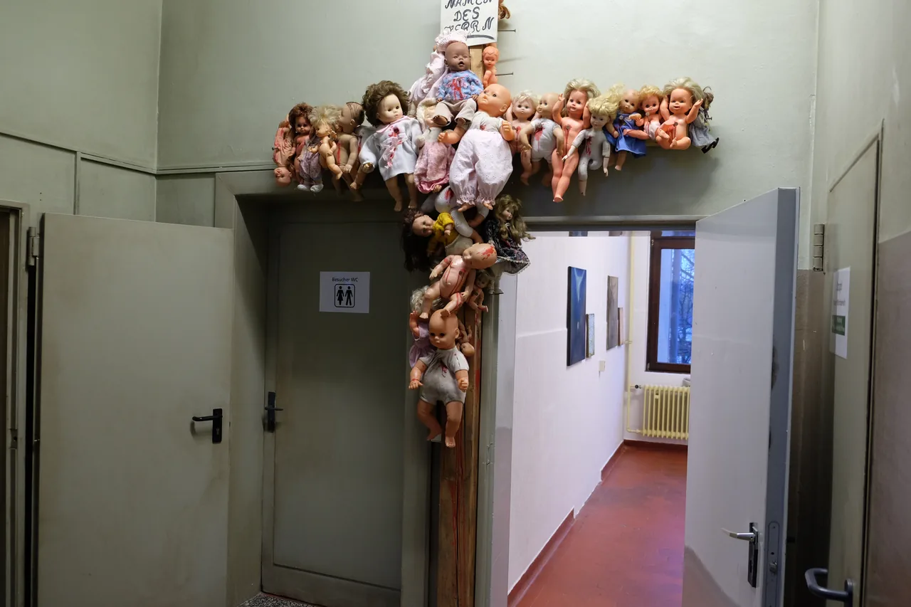 Das Kreuz mit den Puppen hängt in einem Flur über einer Tür.