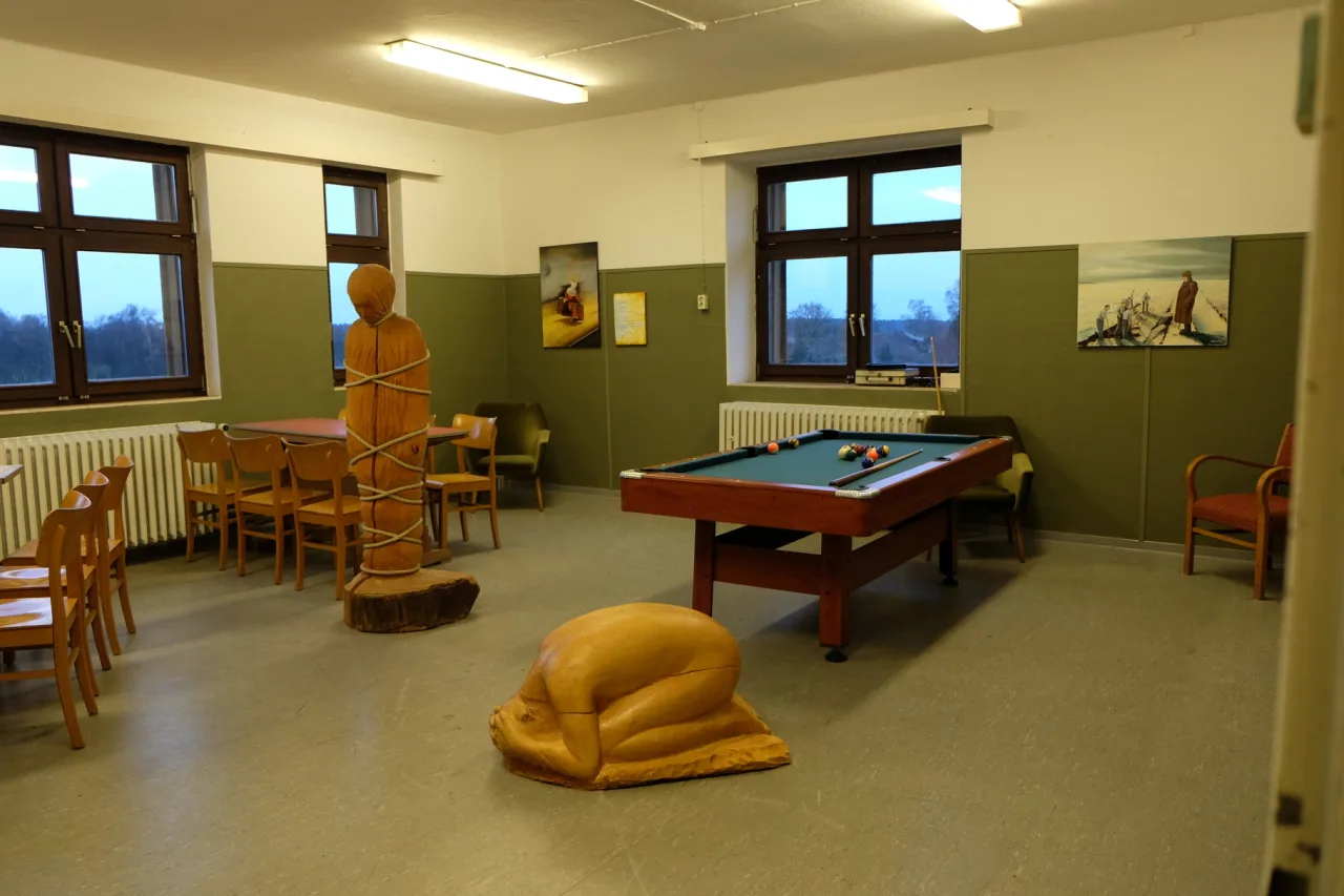 Ein Raum mit Billardtisch und Stühlen, darin eine mit Seilen umwickelte Holzfigur und eine kauernde Holzfigur, an den Wänden Bilder.