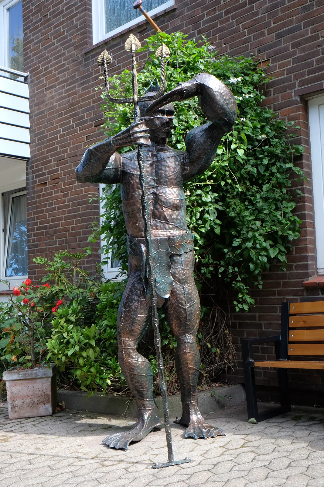 Eine Figur aus Metall, größer als ein Mensch, mit Dreizack und Schwimmflossen vor einer Backsteinwand; ein Arm endet in einer Hummerschere.