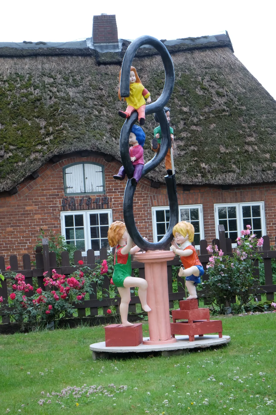 Mehrere farbig gefasste Kinderfiguren an einer geschwungenen dunklen Form über einem rosa Sockel - Eckhard Kowalke